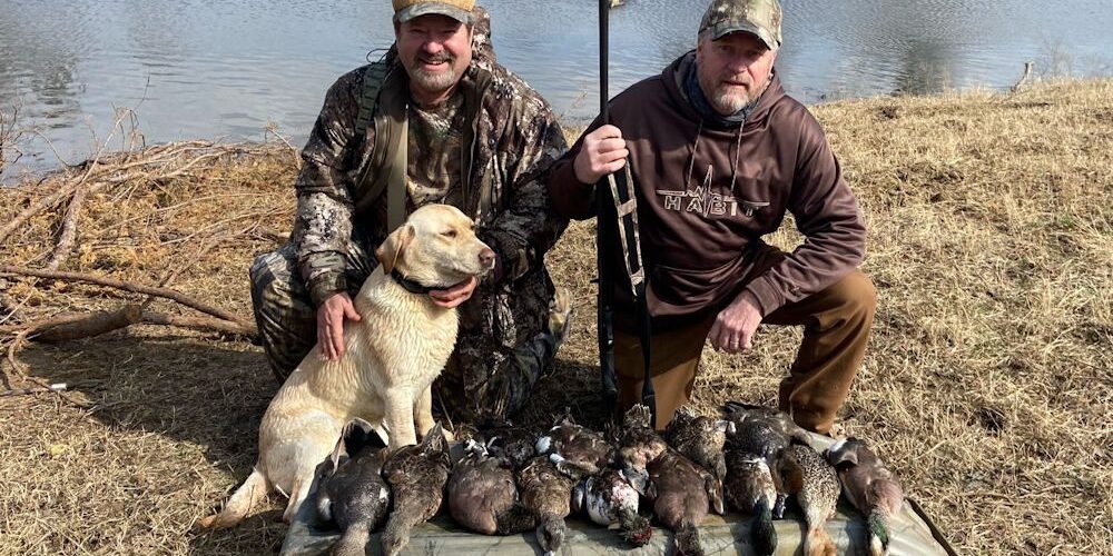 Two hunters by pond with dog and ducks Two hunters by pond with dog and ducks