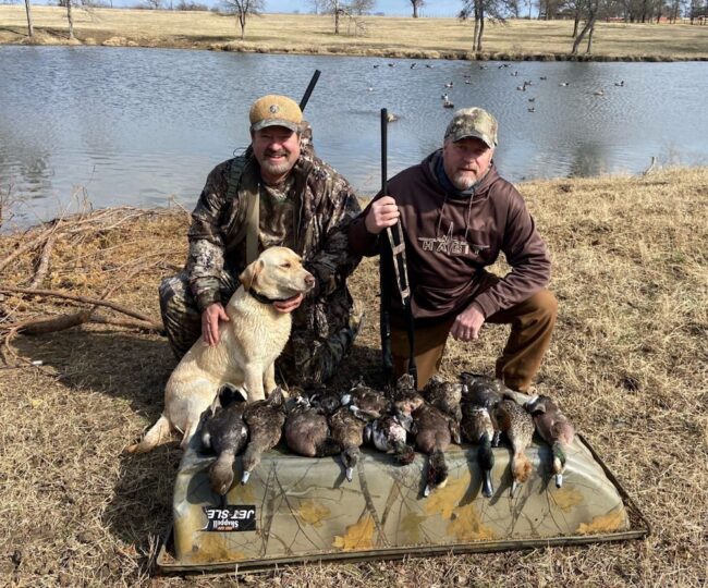 Two hunters by pond with dog and ducks